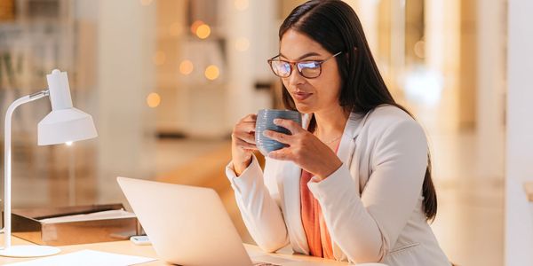 Woman holding a mug while sitting at a desk and looking at an open laptop.