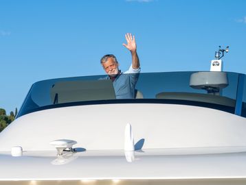 Man waving from a yacht's cockpit with a woman beside him under a clear sky.