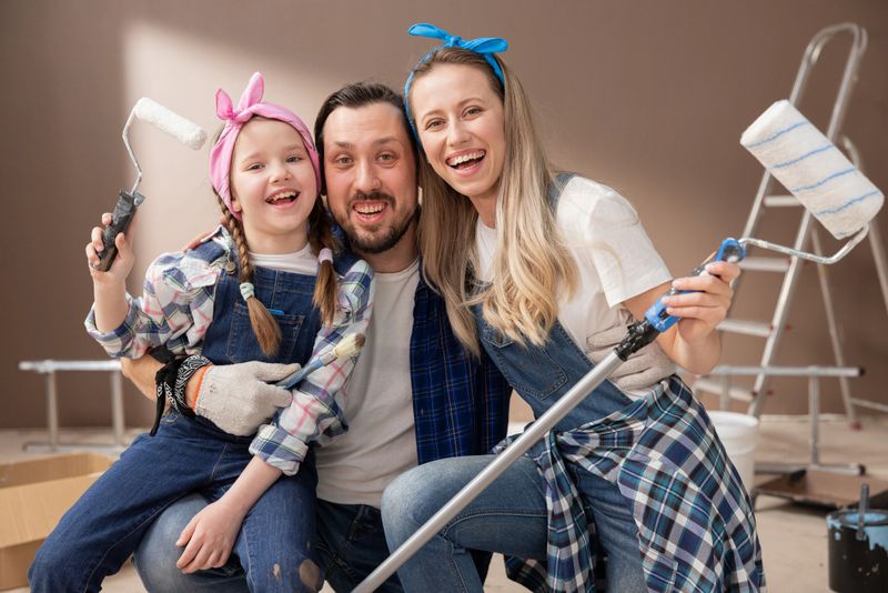A loving family huddles while renovating an apartment. They hold paint rollers to paint the walls. The child and parents smile at how happy they are with the move.