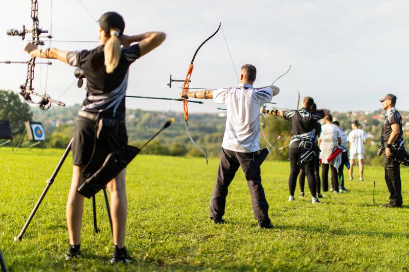Women and men practicing archery training with recurve and compound bow on open field before sunset.