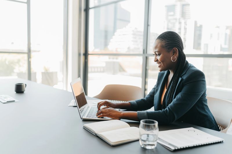 Happy business woman typing on laptop in an office doing market research. Professional female corporate worker with a smile reading an online email or funny social media meme in a startup company