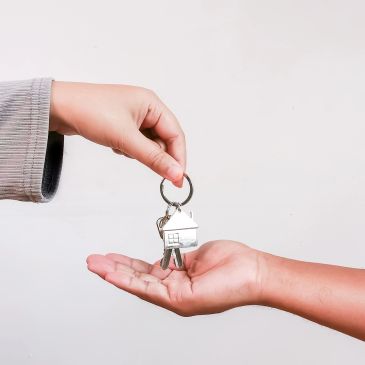 Hand passing house-shaped keychain keys to another hand.