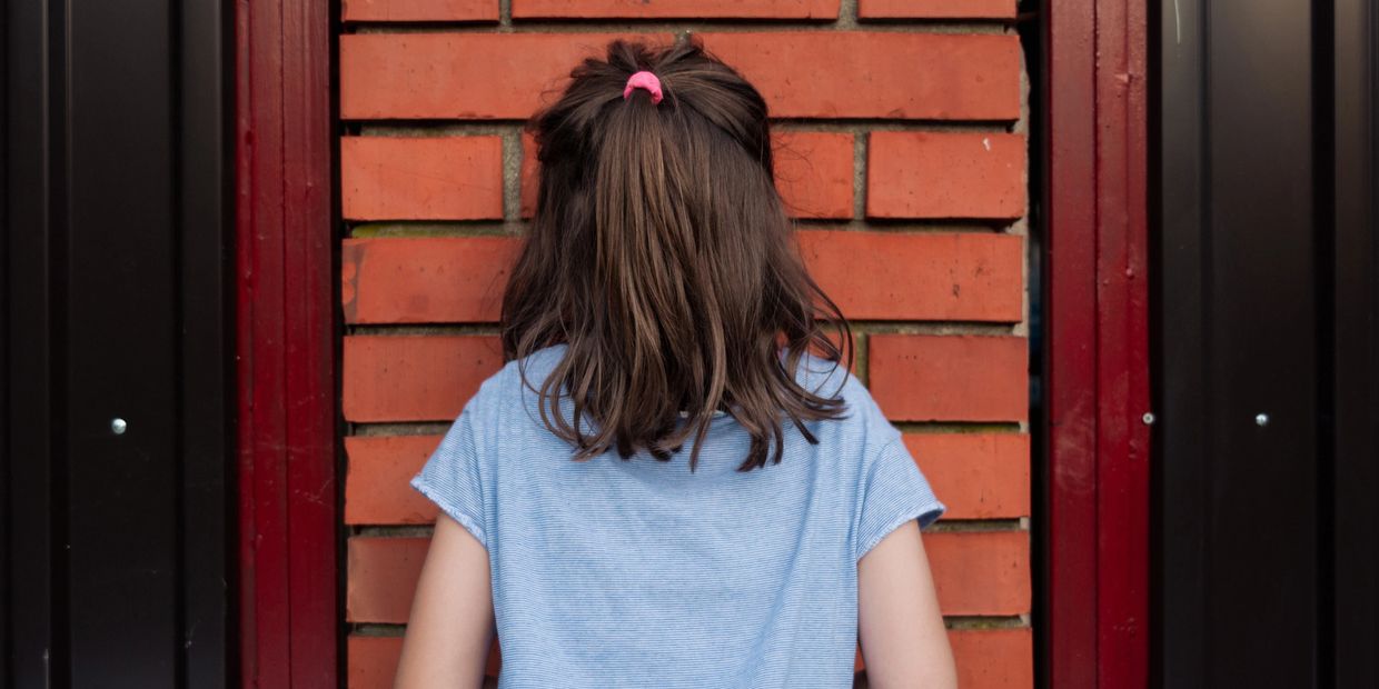 A girl with a pink hair tie stands facing a brick wall.
