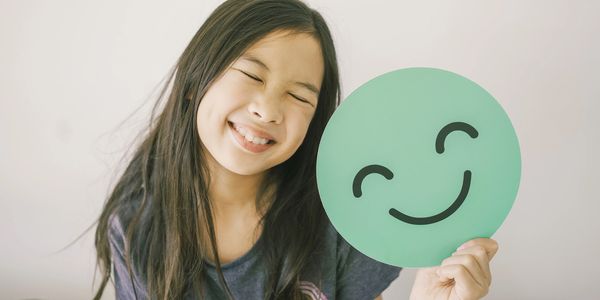 Young girl smiling with her eyes closed holing up a round smiley face symbol next to her head