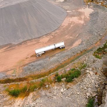 A tanker truck parked on a dirt road near a large gravel pile in a quarry.