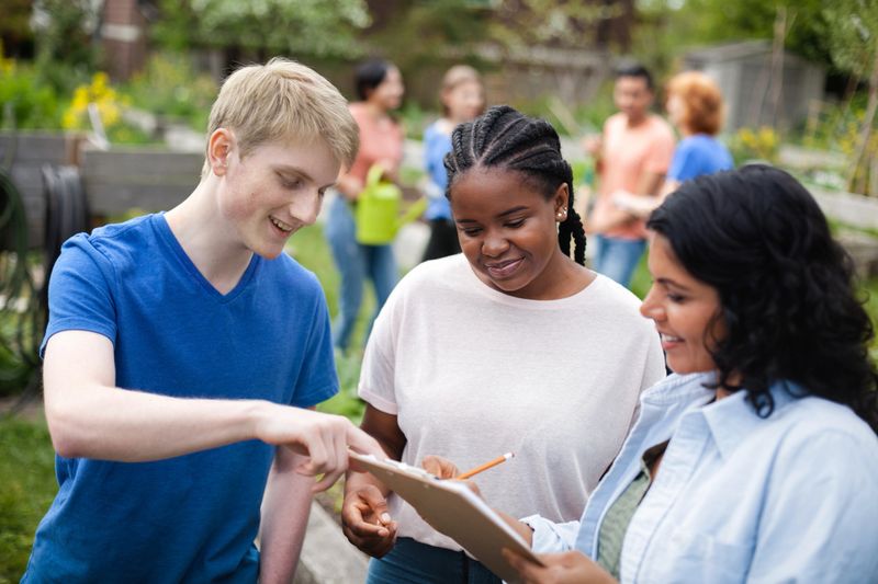 Diverse multiracial group of young volunteers coordinate plans together with mature project manager at community garden park in neighborhood environment