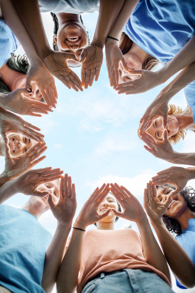 Multiracial group of diverse people stand in circle as community volunteers to show support and commitment to teamwork success togetherness making hand gesture in concept symbol sign of heart shape outdoors with sky