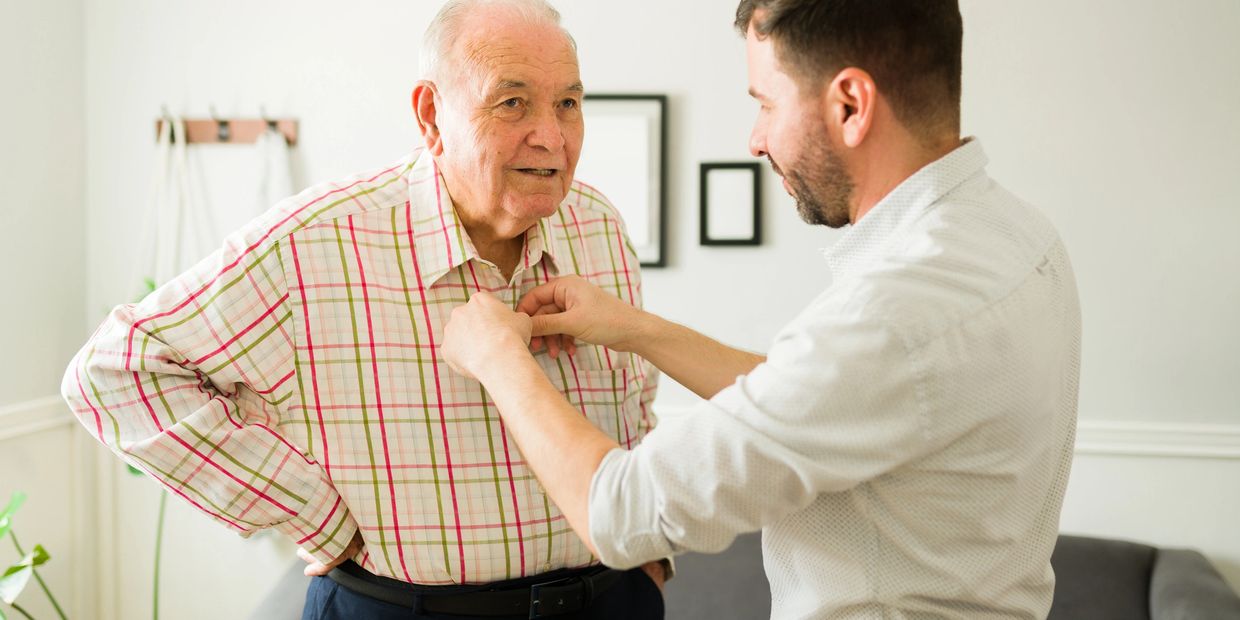 A younger man helps an elderly man button his shirt indoors.