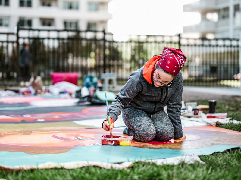 Young Latin woman creating outdoor mural on the lawn. She is dressed in casual outfit, wearing bandana and eyeglasses. Exterior of public park in the city.
