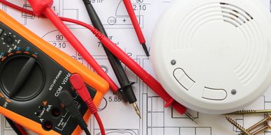 Digital multimeter and smoke detector on blueprints for fire alarm installation and testing