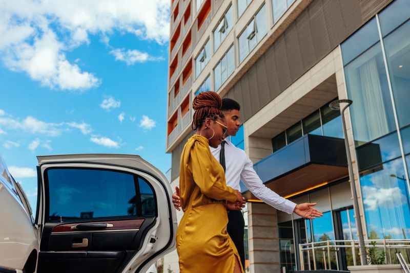 A young valet is escorting a wealthy female African American client to the hotel out of the limousine.