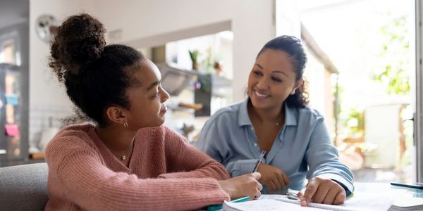 A woman helps a teenage girl study at a table with notebooks and pens.