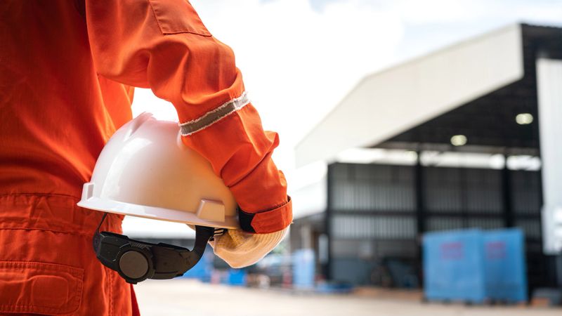 An operation worker in orange coverall is holding white safety helmet or hardhat with factory place as background. Safe working practice in the industrial scene photo, close-up and selective focus.