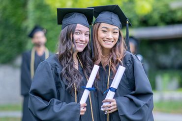 Two happy female graduates in caps and gowns holding diplomas outdoors.