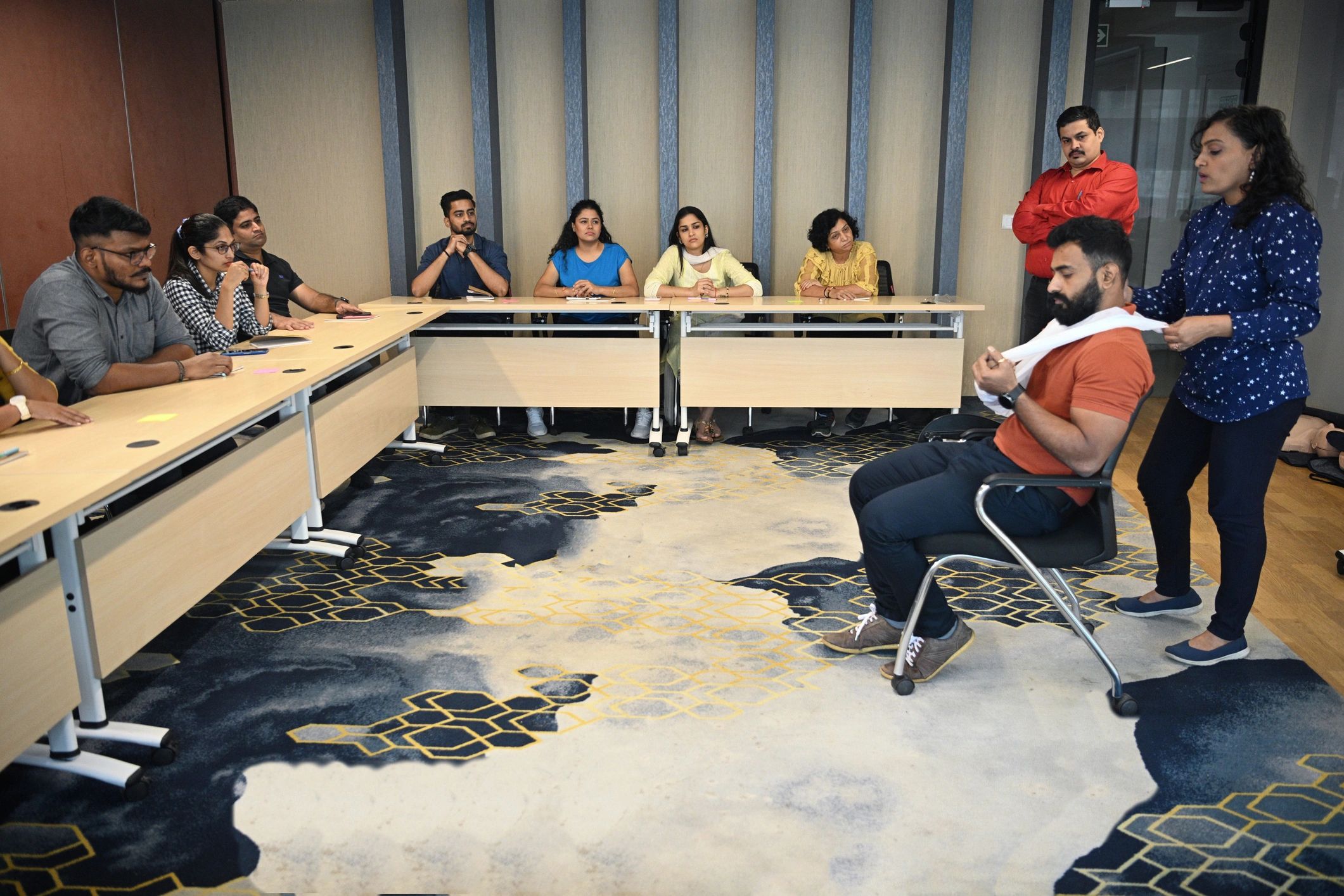 A woman demonstrates a first aid technique on a seated man in a meeting room.