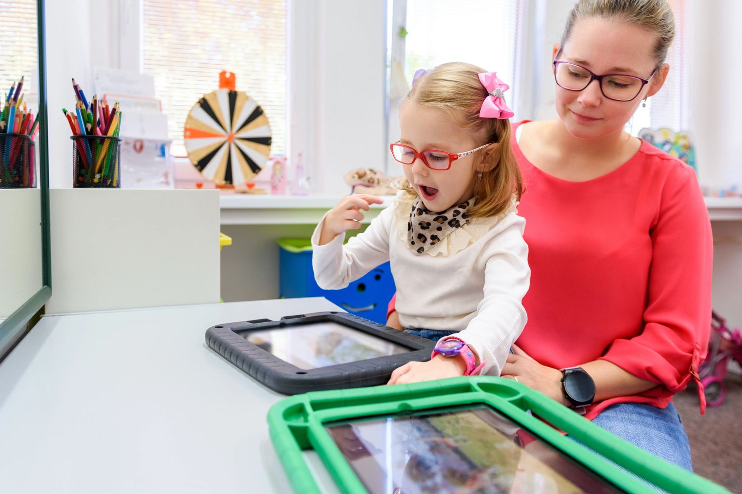 Young girl with glasses using a tablet, guided by a woman in a classroom.