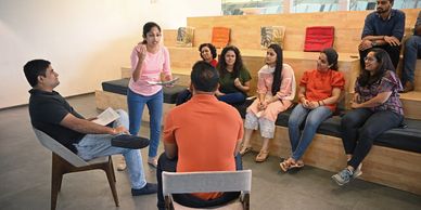 A woman in a pink shirt speaks to a group seated in a modern meeting space.