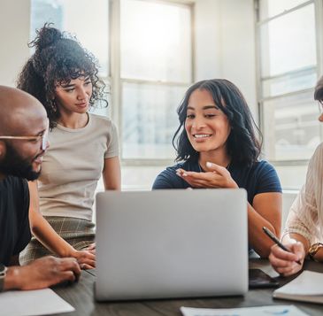 Four colleagues discuss work around a laptop in a bright office.