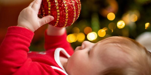 Baby in red holding a Christmas ornament with festive lights in the background.