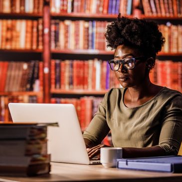 Woman studying on laptop in a cozy library setting with books and a lamp.