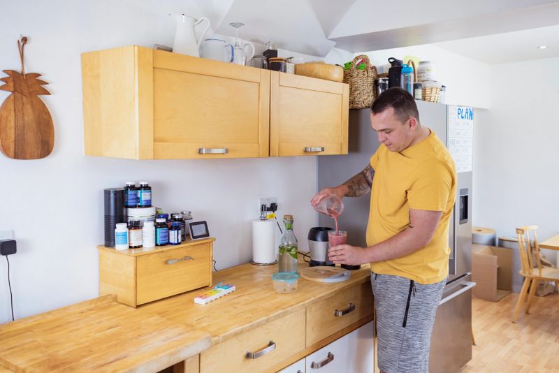 Man standing in his kitchen in the North East of England making a smoothie. He is using fresh fruits, eating healthily as part of his new fitness plan. He is adding supplements to the juicer.