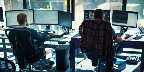 Two software developers working at desks with multiple monitors in a sunlit office.
