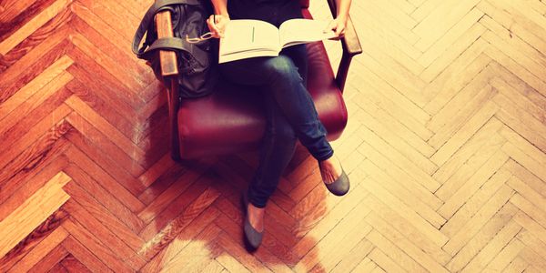 Person sitting on a chair reading a book with a backpack beside them.