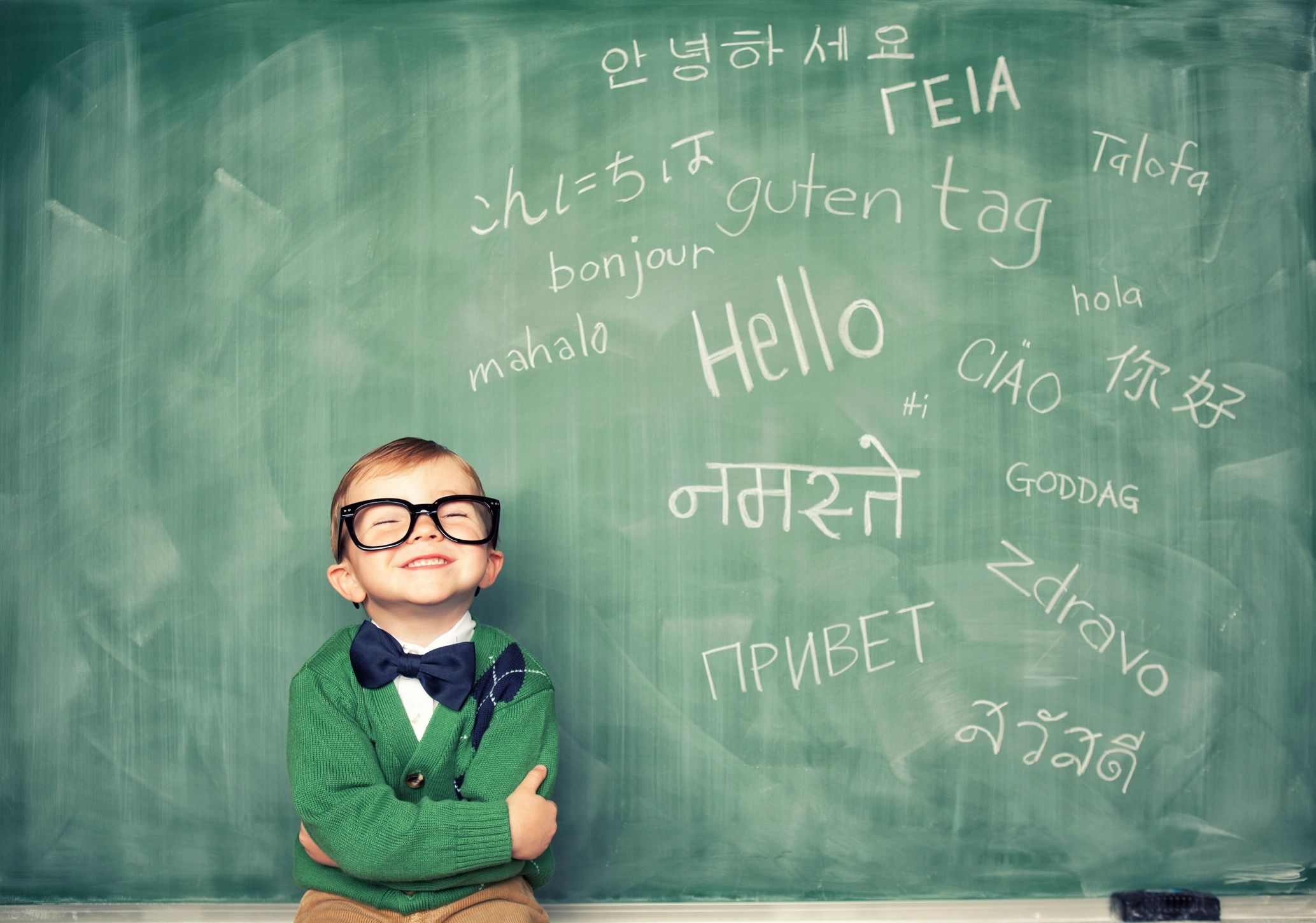 Smiling child in glasses and bow tie with multilingual greetings on chalkboard.