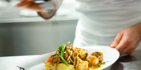 Chef garnishes a plated gourmet dish with sauce in a professional kitchen.