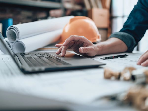 Engineer working on laptop with blueprints and helmet.