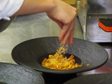 Chef garnishing a pasta dish with grated cheese in a kitchen.