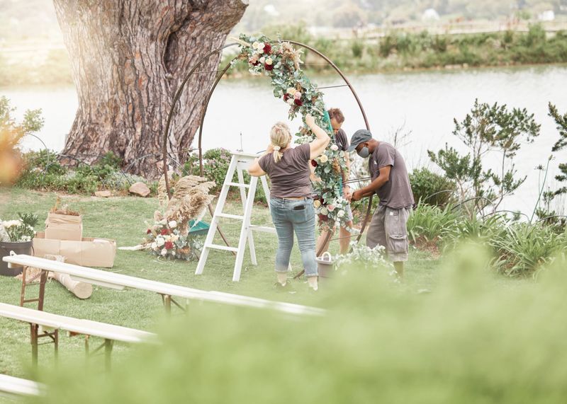 Wedding ceremony, florist or event planner decorating arch with flowers for floral theme by quiet, calm and beautiful lake with team. Decoration, marriage and romance with decor staff for celebration