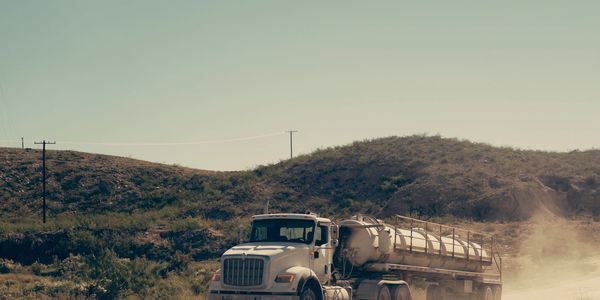 A tanker truck driving on a dusty road in a hilly, dry landscape.
