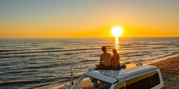 A couple sits on a van roof watching a sunset by the beach.