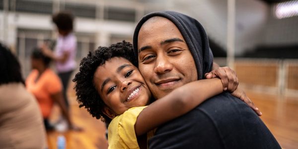 Smiling child hugs a man in a hoodie at an indoor sports court.