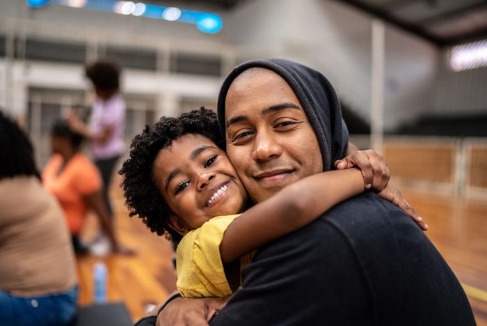 Smiling child hugs a man in a hoodie at an indoor sports court.