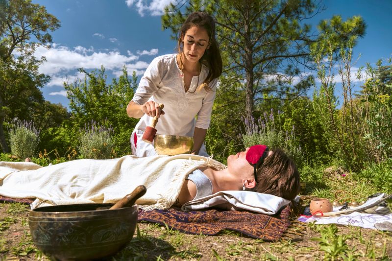 Woman therapist practicing sound healing therapy on a woman lying in a natural environment.