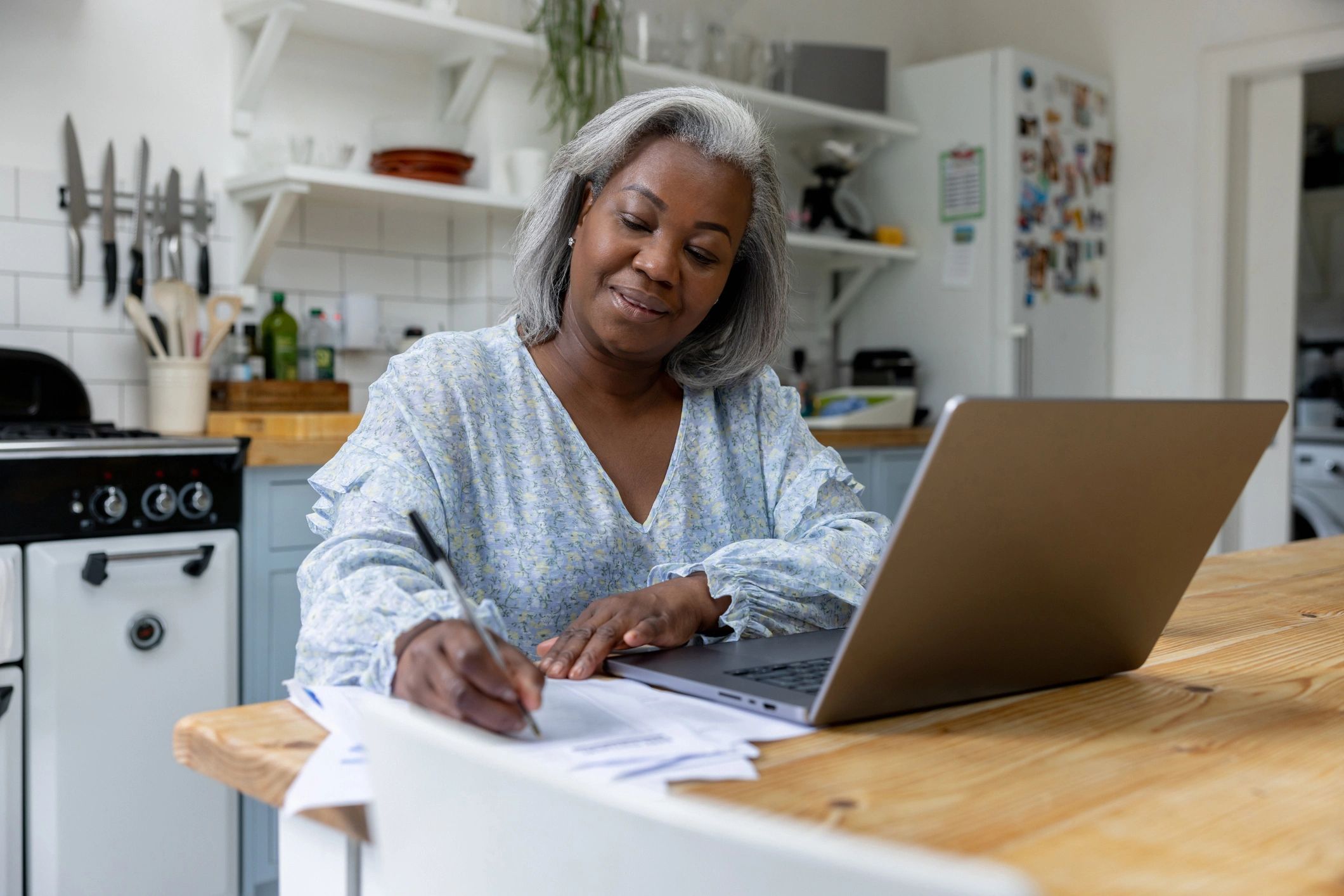 A woman writes notes while working on a laptop in her kitchen.