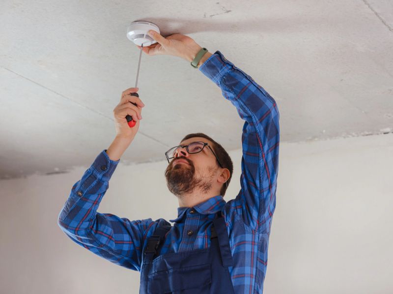 young electrician man in blue work suit installing smoke detector on the ceiling