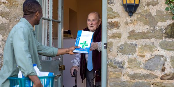 Man delivering medicine to elderly man using a walker at his doorstep.