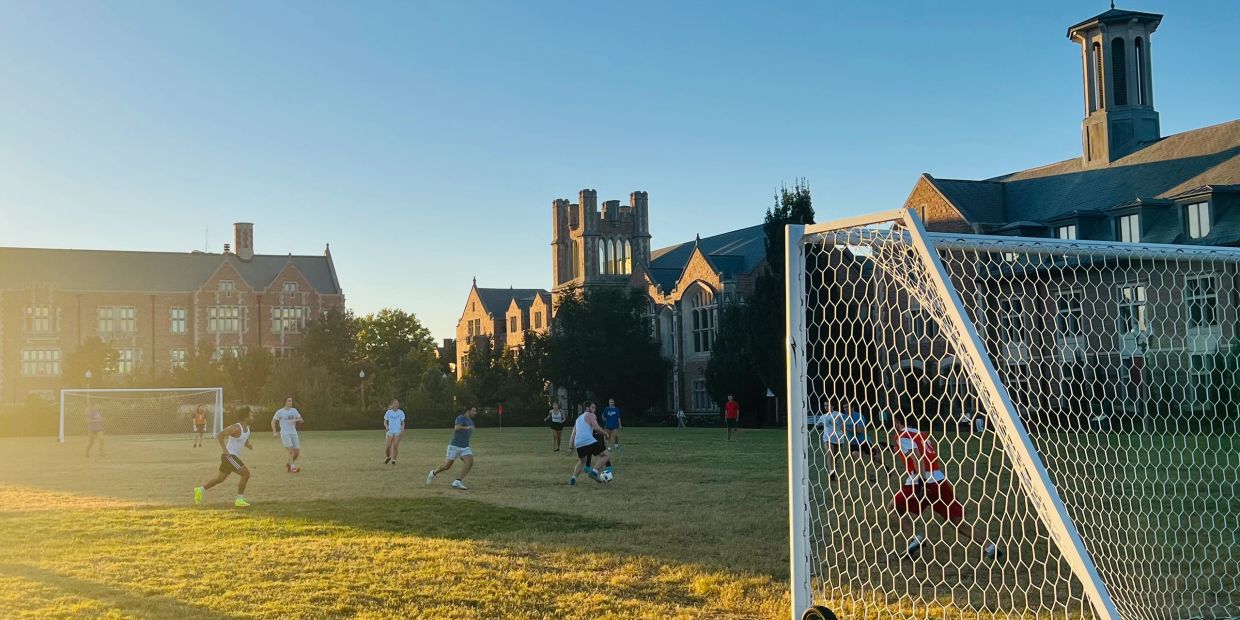 People playing soccer on a grassy field with historic buildings in the background at sunset.