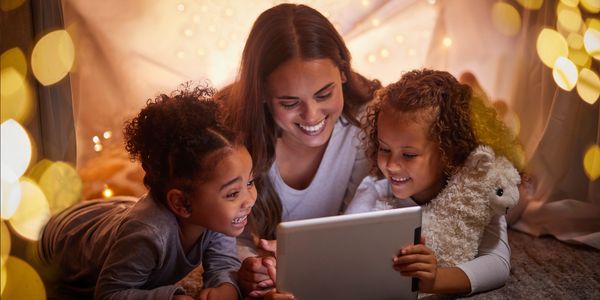 A woman and two children happily looking at a tablet inside a cozy fort.