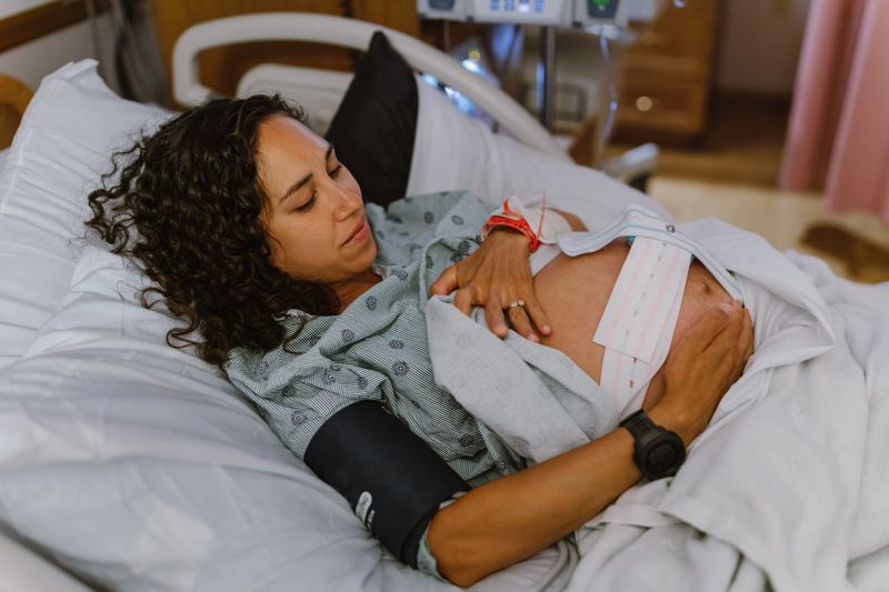 A beautiful Eurasian pregnant woman lies in a hospital bed and smiles lovingly down toward her abdomen in anticipation of meeting her newborn baby.