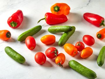 Fresh mini cucumbers, cherry tomatoes, and mini bell peppers on a white surface.