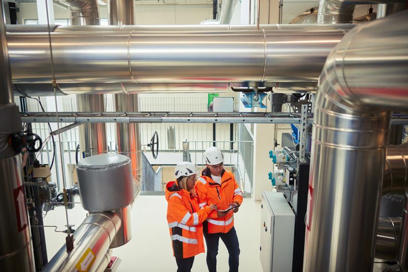 Two engineers working with a digital tablet while standing by a control panel in a station producing heat for homes using water