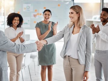 Colleagues in an office smiling and shaking hands while others applaud.