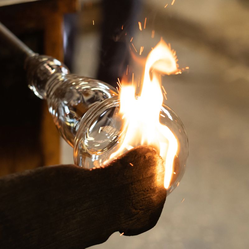 The process of glassblowing begins with taking a blob of molten glass with a blowpipe, rotating it to give a first shape, and then  blowing into it to create a bubble. Then the glassblower rotates it and shapes it with his tools, ensuring it stays at the right temperature by putting it again rapidly in the furnace. At the end of the process, the object is attached to a pontil so that it can be taken off the blowpipe to shape the aperture.