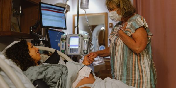 Pregnant woman in hospital bed holds hands with a masked visitor.