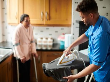 Caregiver ironing clothes while elderly woman with crutches watches in kitchen.