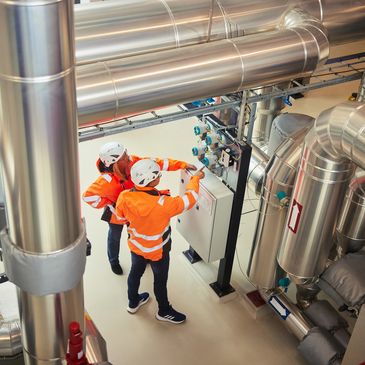 Two workers in orange safety jackets inspecting industrial machinery with pipes.